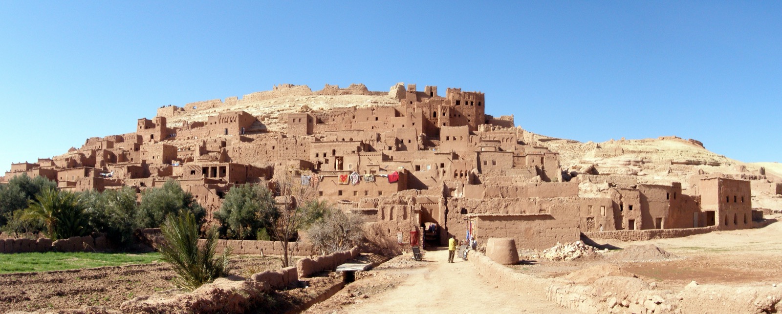 Panoramablick auf den Ksar von Ait Ben Haddou in Marokko: ein befestigtes Lehmbaudorf aus dicht gedraengten Stampflehm- und Lehmziegelhäusern, das sich terrassenfoermig den Huegel hinaufzieht, unter klarem blauem Himmel.